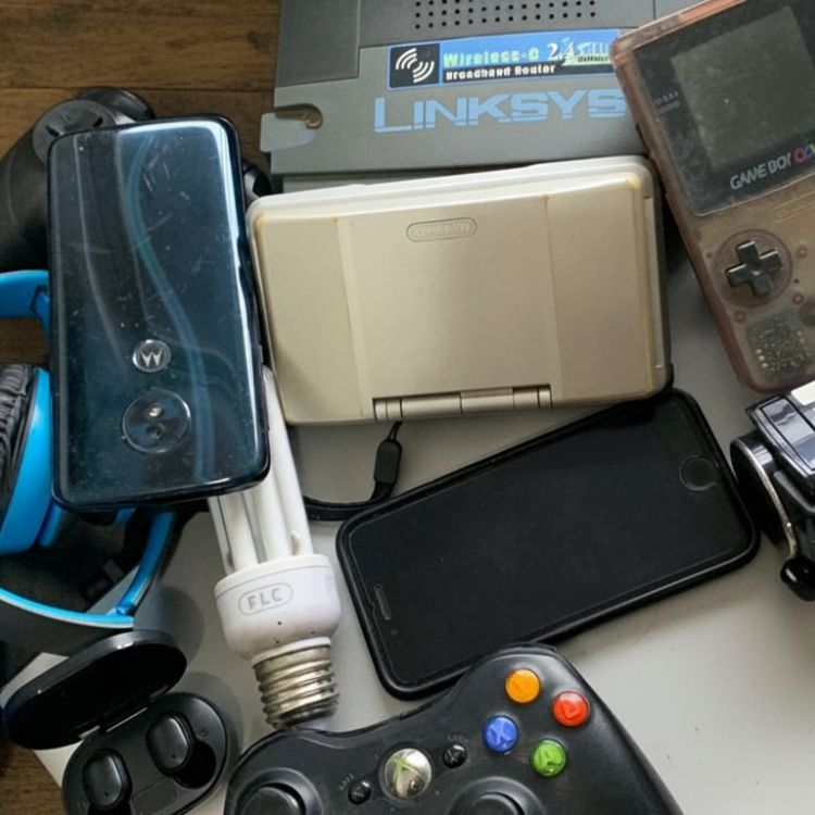 Pilha de eletrônicos antigos e quebrados, como celulares e notebooks, em centro de reciclagem com símbolo de reciclagem ao fundo